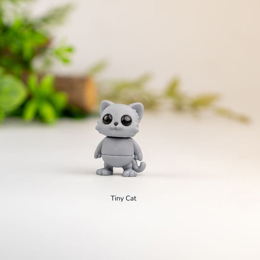 Small gray cat figurine on a white surface with green and orange flowers in the background