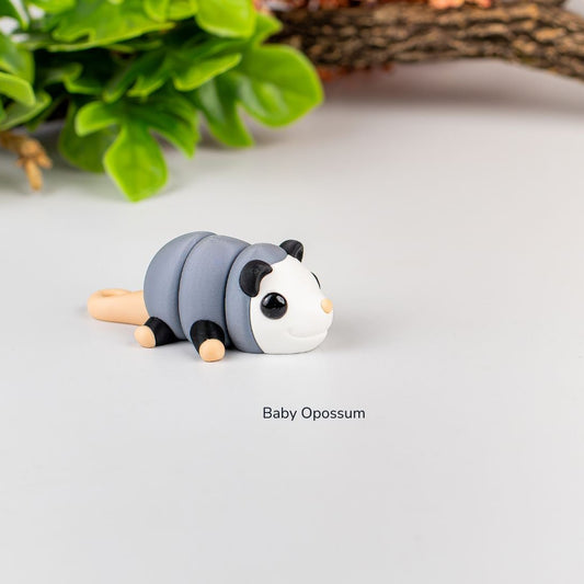 Tiny baby opossum Pocket Zoo figure with a segmented gray body, white face, black ears and feet, and a pale peach tail.