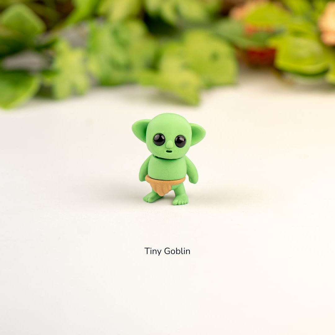 Small green figurine labeled 'Tiny Goblin' on a white background with blurred greenery in the background.