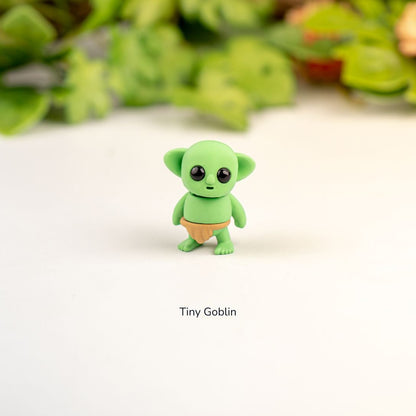 Small green figurine labeled 'Tiny Goblin' on a white background with blurred greenery in the background.