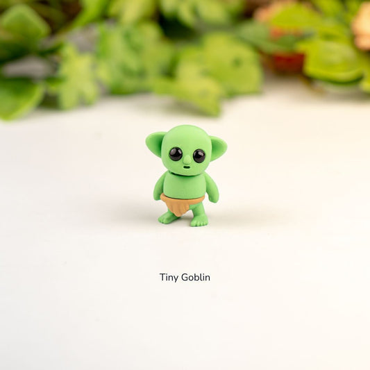 Small green figurine labeled 'Tiny Goblin' on a white background with blurred greenery in the background.